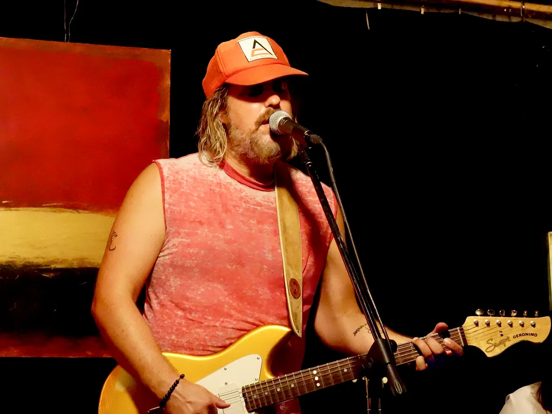 Brad Byrd singing and playing a gold electric guitar beside a large red and gold painting at The Garage, an art and performance space